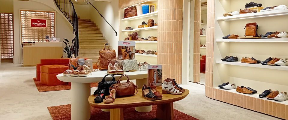 Interior of a Pikolinos shop displaying sandals and bags on a round table in the foreground, with shelves full of assorted footwear in the background and a staircase leading to the upper floor.