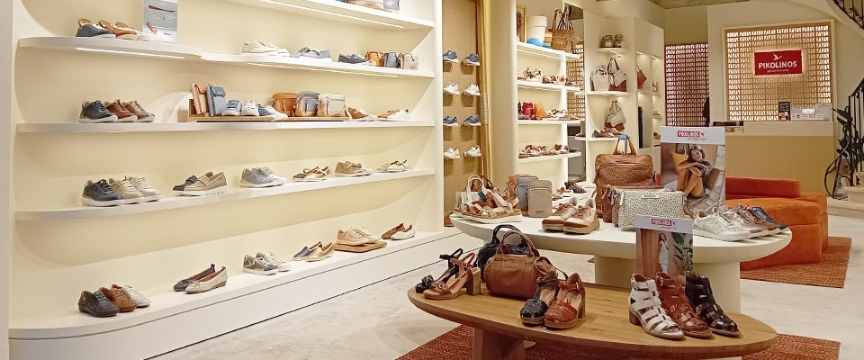 Interior of a Pikolinos shoe shop with illuminated shelves full of shoes and a central table displaying sandals and leather bags in natural tones.