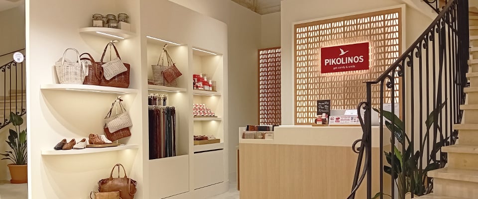 Interior of a Pikolinos shop with illuminated shelves displaying bags, footwear and accessories. On the right, a counter with a Pikolinos logo sign and a spiral staircase with a wrought iron railing.